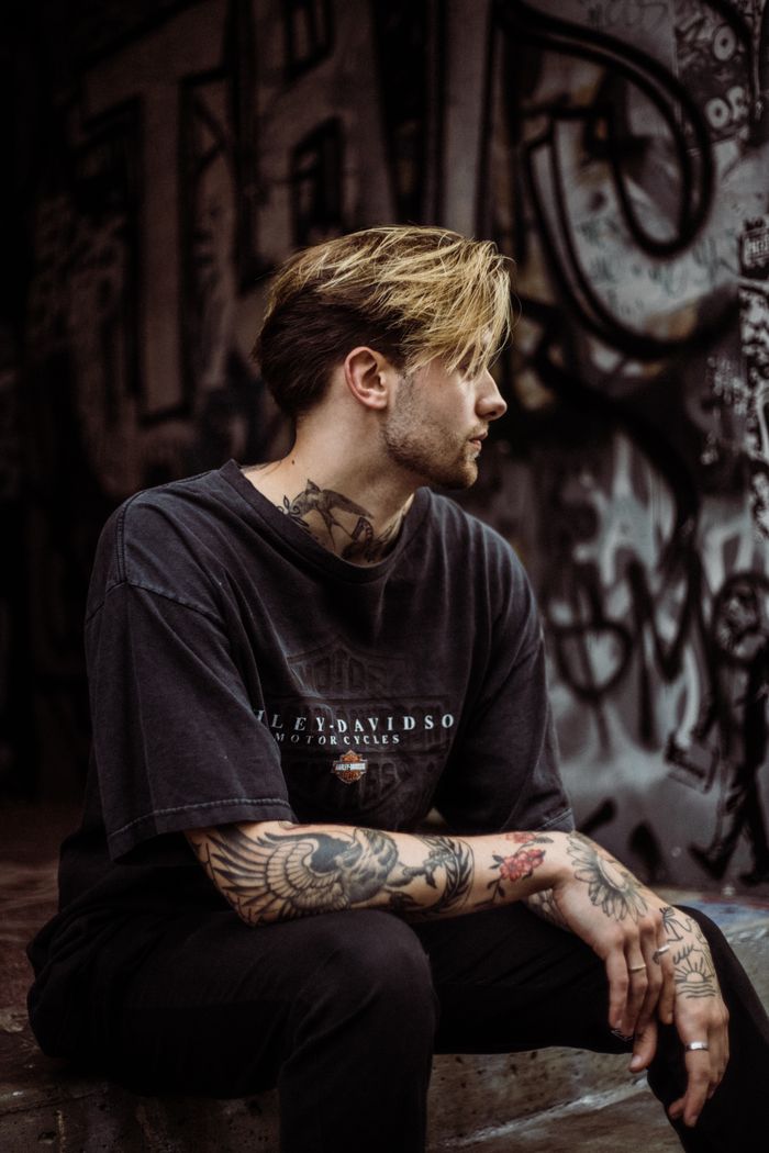 who-we-are A tattooed man in a black T-shirt sits in a graffiti-covered urban environment, showcasing street style.
